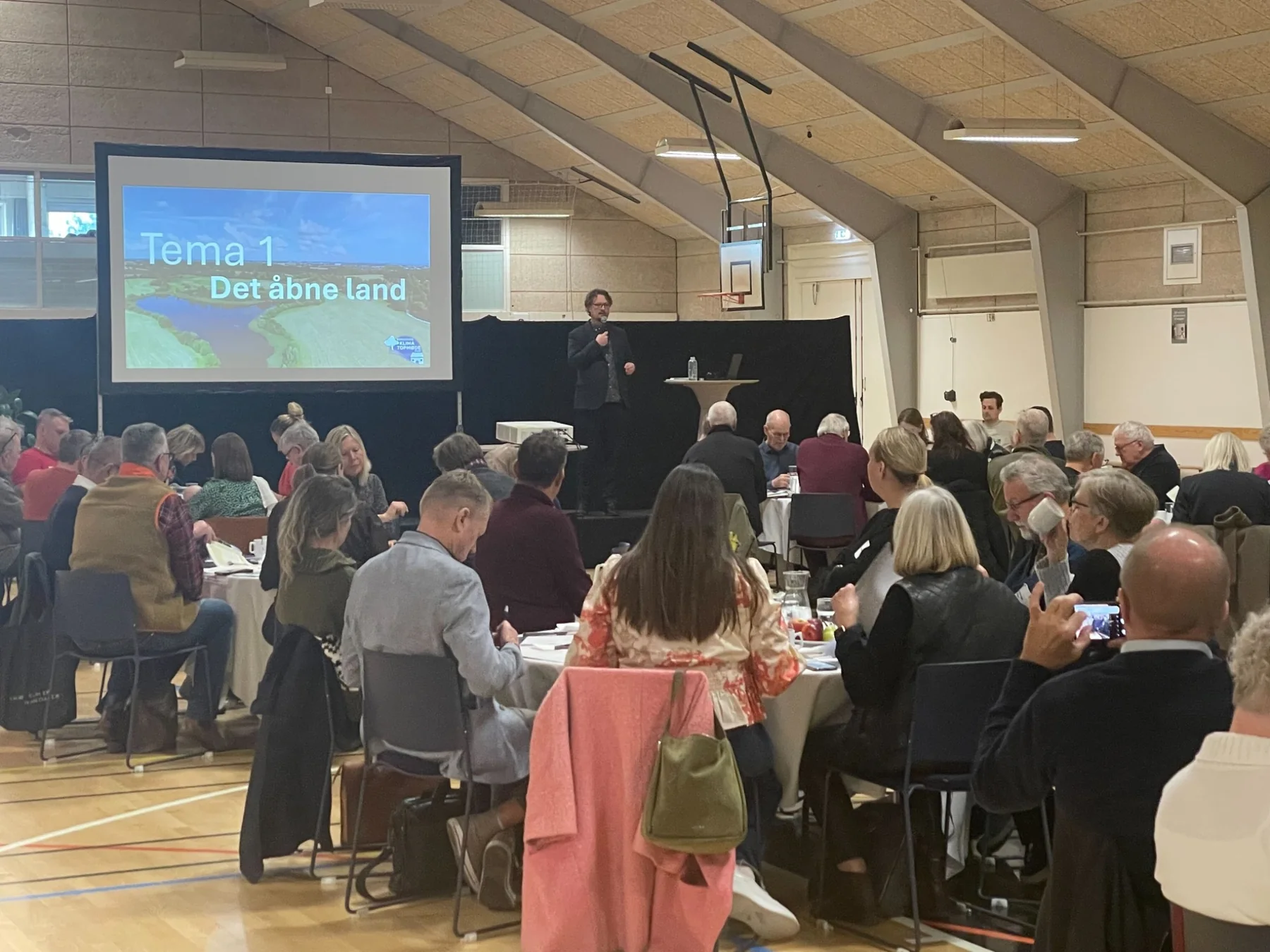Mark holding a microphone in front of participants gathered around tables in a community hall